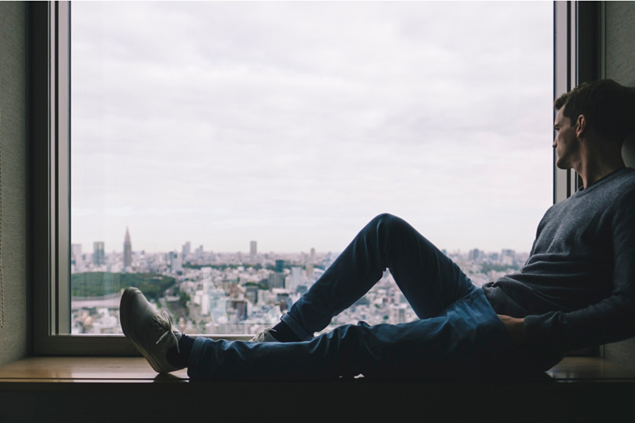 Image d'en-tête de l'article Je Réalise " 12 erreurs de pensée alimentant nos émotions désagréables ", sur laquelle l'on voit un homme assis dans le large encadrement d'une fenêtre, les mains dans les poches, regardant la ville par la fenêtre.
