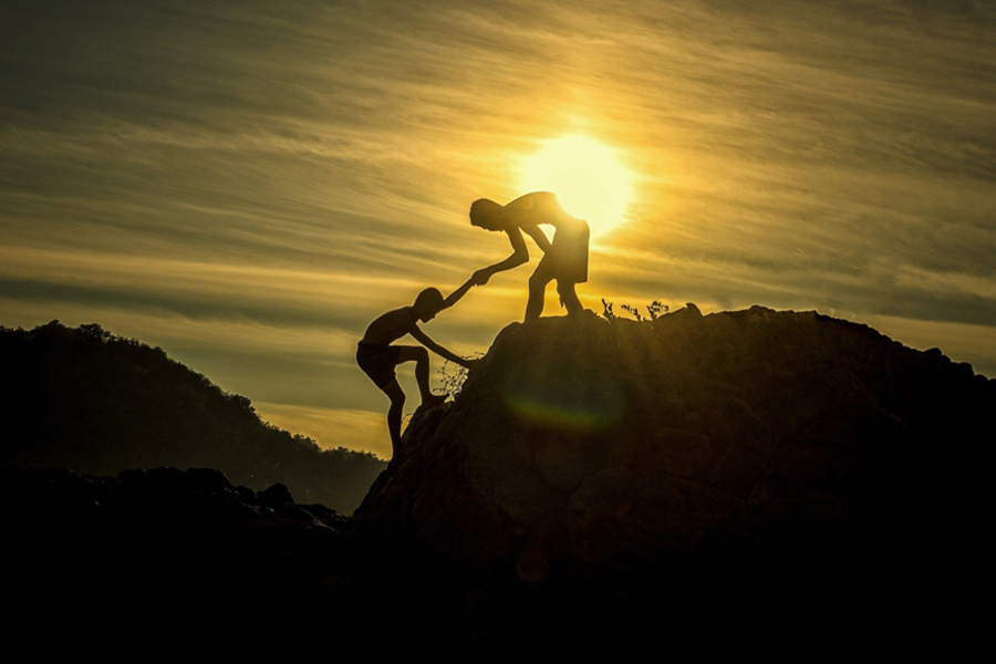 Image d'en-tête de l'article Je Réalise " Aider " oui ", mais sans me sacrifier ", sur laquelle l'on voit un homme au sommet d'une montagne au soleil couchant, prendre la main d'un enfant pour l'aider à monter.