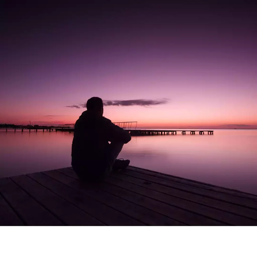 Image d'en-tête de l'article Je Réalise : " Apprivoiser nos émotions " ", sur laquelle l'on voit une silhouette d'homme assit sur un ponton au bord de l'eau contemplant les belles couleurs du crépuscule.
