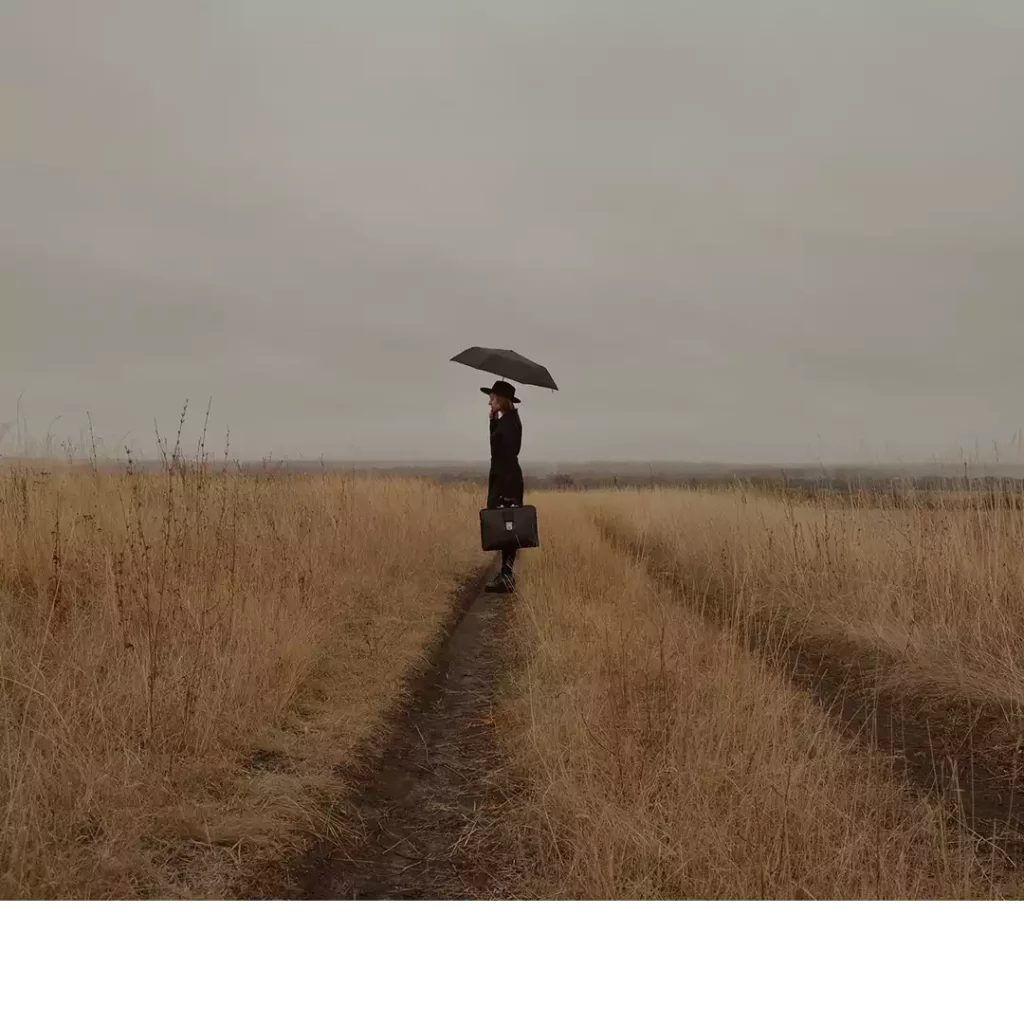 Image d'en-tête de l'article Je Réalise : " Apaiser la tristesse, retrouver le sourire ", sur laquelle l'on voit une personne tournée vers la gauche, seule dans un champ, tenant un parapluie sous un ciel gris.