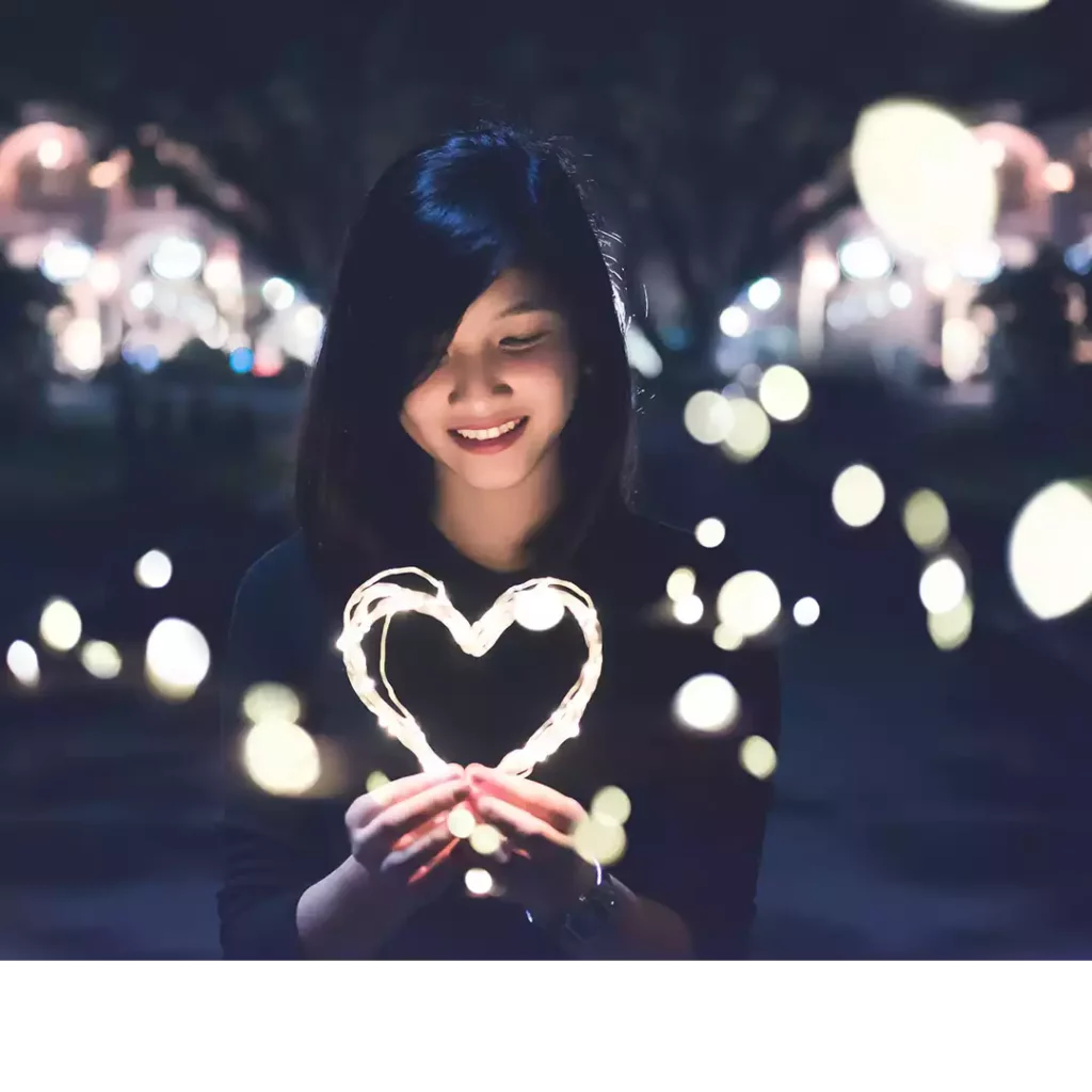 Image d'en-tête de l'article Je Réalise : " Et si le grand amour commençait par toi ? 5 exercices pour apprendre à t’aimer ", sur laquelle l'on voit une femme souriante tenant dans ses mains un cœur lumineux et rayonnant symbolisant l'amour de soi.